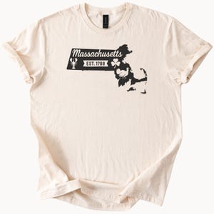 Könnte beinhalten: Cremefarbenes T-Shirt mit einer schwarzen Grafik der Umrisse des Bundesstaates Massachusetts. Das Design beinhaltet ein Banner mit dem Wort "Massachusetts" und "EST. 1788" sowie eine Silhouette von Hummer, Kleeblatt und Truthahn.