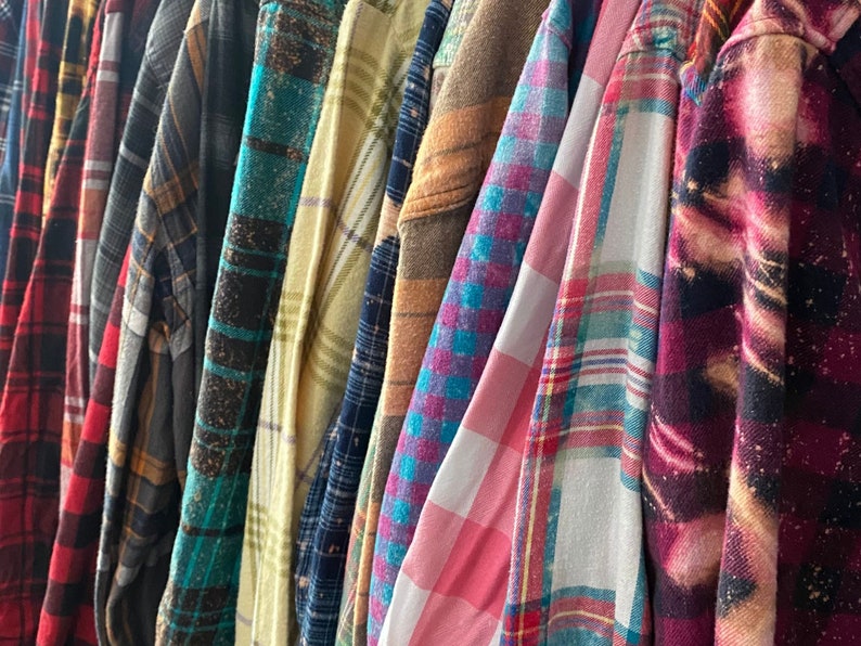 May include: A collection of plaid flannel shirts in various colors and patterns, including red, blue, green, yellow, and brown. The shirts are hanging on a rack.
