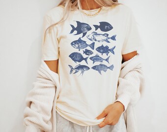 May include: A cream colored t-shirt with a blue graphic of various fish. The fish are all facing different directions and are arranged in a cluster.