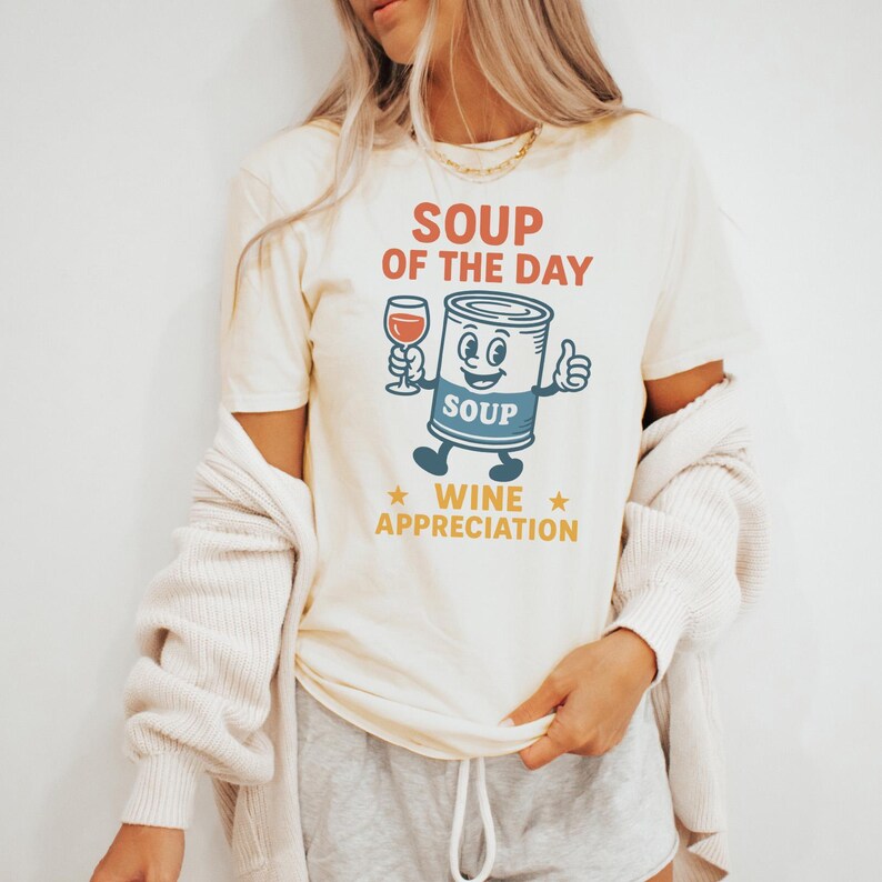 Op de afbeelding: Cr&egrave;mekleurig T-shirt met een cartoon soepblik karakter dat een wijnglas vasthoudt en een duim omhoog geeft. De tekst op het shirt luidt "SOUP OF THE DAY" en "WINE APPRECIATION" in rood en goud.
