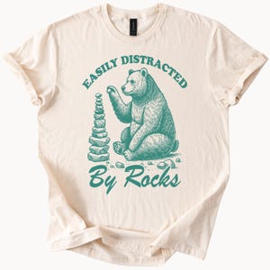May include: Cream-colored t-shirt with a teal graphic of a bear stacking rocks. The text "EASILY DISTRACTED" arches above the bear, and "By Rocks" is below. The bear is detailed with shading, and the rocks are stacked in a tower.
