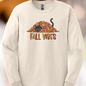 May include: Cream-colored long-sleeve shirt featuring a black cat peeking out from a pile of autumn leaves. The phrase "FALL VIBES" is printed in orange, yellow, and red. The shirt is a casual, seasonal apparel item.