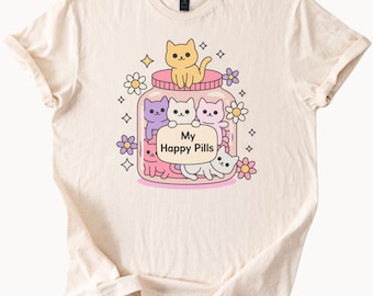 Könnte beinhalten: Cremefarbenes T-Shirt mit einer skurrilen Illustration von Cartoon-Katzen in einem Glas mit der Aufschrift "My Happy Pills". Das Design umfasst verschiedenfarbige Katzen, Blumen und funkelnde Akzente, die eine verspielte Ästhetik erzeugen.