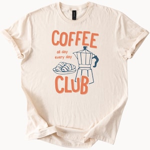 May include: Cream-colored t-shirt with the words "COFFEE CLUB" in orange, with "all day every day" in between. A blue and white coffee maker and a plate of croissants are also featured.