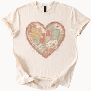 Peut inclure: T-shirt crème avec un motif en forme de cœur composé de divers motifs patchwork aux couleurs douces comme le pêche, le vert sauge et le beige. Le cœur a une bordure effilochée et un aspect légèrement vieilli.