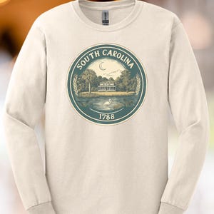 Puede incluir: Camiseta de manga larga color crema con un gráfico circular del sello del estado de Carolina del Sur. El sello incluye una casa, árboles, una luna creciente y el texto "SOUTH CAROLINA" y "1788" en verde azulado.