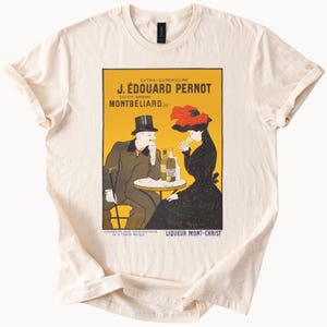 Puede incluir: Camiseta color crema con un anuncio vintage de licor J. Édouard Pernot. El diseño incluye una pareja en una mesa, botellas y texto en francés. La camiseta tiene cuello redondo y mangas cortas.