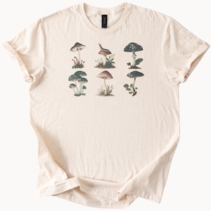 May include: Cream-colored t-shirt featuring a botanical illustration of various mushrooms. The design includes detailed drawings of different mushroom species in shades of green, brown, and red. The shirt has a classic crew neck and short sleeves.