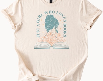 May include: A beige t-shirt featuring a graphic design of a woman with her back to the viewer, reading a book. The text "Just a girl who loves books" is printed around the woman's head.