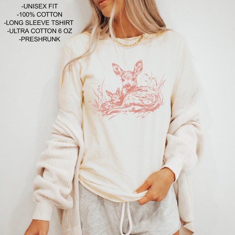 May include: A cream-coloured long-sleeve t-shirt featuring a pink illustration of a deer and fawn. The shirt is made of 100% cotton and is pre-shrunk. The text on the shirt reads "-UNISEX FIT -100% COTTON -LONG SLEEVE TSHIRT -ULTRA COTTON 6 OZ -PRESHRUNK".