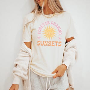 May include: A white t-shirt with a pink and yellow graphic design that says "Forever Chasing Sunsets" with a stylised sun and two stars.