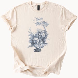 May include: Cream-colored t-shirt featuring a blue and white pastoral scene. The graphic depicts figures under a tree, with detailed foliage and a serene atmosphere. The shirt has a classic crew neck and rolled sleeves.