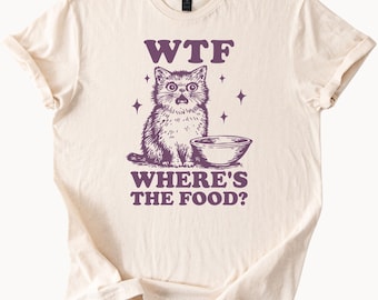 May include: Cream-colored t-shirt with a purple graphic of a surprised cat and a food bowl. The text "WTF WHERE'S THE FOOD?" is printed above and below the cat illustration. The shirt has short sleeves and a crew neck.