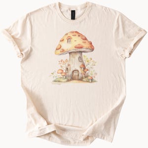 May include: Cream-colored t-shirt featuring a watercolor illustration of a mushroom house. The mushroom has a brown cap with orange spots and a door and windows. Surrounding the mushroom are smaller mushrooms and foliage.