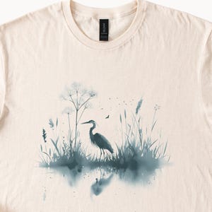 May include: A watercolor illustration of a heron standing in a marsh with tall grasses and a blue-grey color scheme.