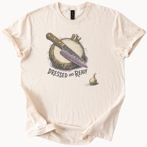 May include: A cream-colored short-sleeve t-shirt featuring a graphic illustration of a chef's knife resting on a sliced onion. Below the onion, the text reads DRESSED AND READY in black capital letters, accompanied by a small whole onion nearby.