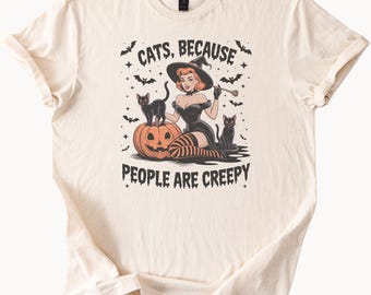 May include: Cream-colored t-shirt featuring a Halloween-themed graphic. The design includes a witch, black cats, a jack-o'-lantern, bats, and the text "CATS, BECAUSE PEOPLE ARE CREEPY". The shirt has short sleeves and a crew neck.