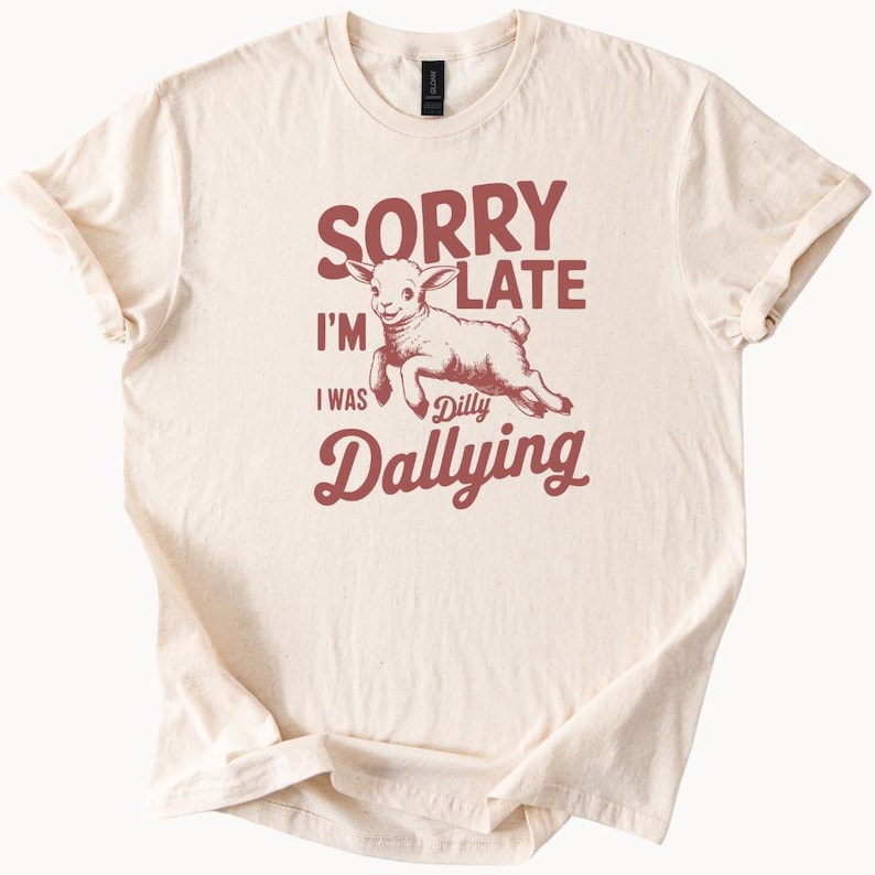 May include: Cream-colored t-shirt with the text "SORRY I'M LATE I WAS Dilly Dallying" in maroon. A cartoon lamb is illustrated in the center, also in maroon. The shirt has short sleeves and a crew neck.