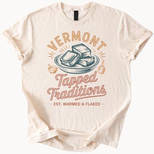 May include: A cream-coloured t-shirt featuring the words "VERMONT" and "Tapped Traditions" in a vintage design. The graphic includes an illustration of maple syrup and the text "EST. WARMED & FLAKED".