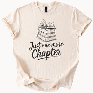 Puede incluir: Una camiseta beige con un gráfico negro de una pila de libros y el texto "Just one more chapter".