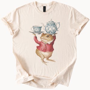 Pode incluir: Camiseta creme com uma ilustração fantasiosa de um sapo de casaco vermelho, segurando um jogo de chá. O jogo de chá apresenta um padrão azul e branco e inclui uma chaleira e uma xícara. O sapo é o foco central do design.