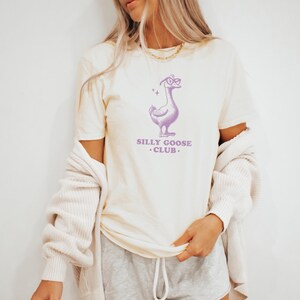 Puede incluir: Una camiseta de color crema con un ganso de dibujos animados morado que lleva gafas y el texto "Silly Goose Club."