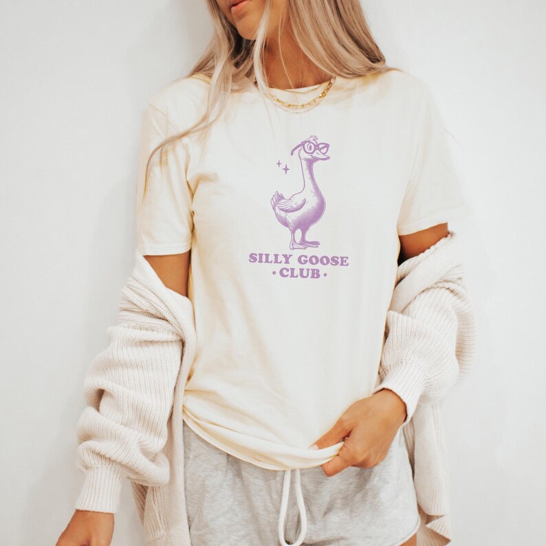 Puede incluir: Una camiseta de color crema con un ganso de dibujos animados morado que lleva gafas y el texto "Silly Goose Club."