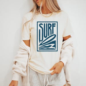 May include: A white t-shirt with a blue graphic of a surfboard and the word "SURF" in a box.