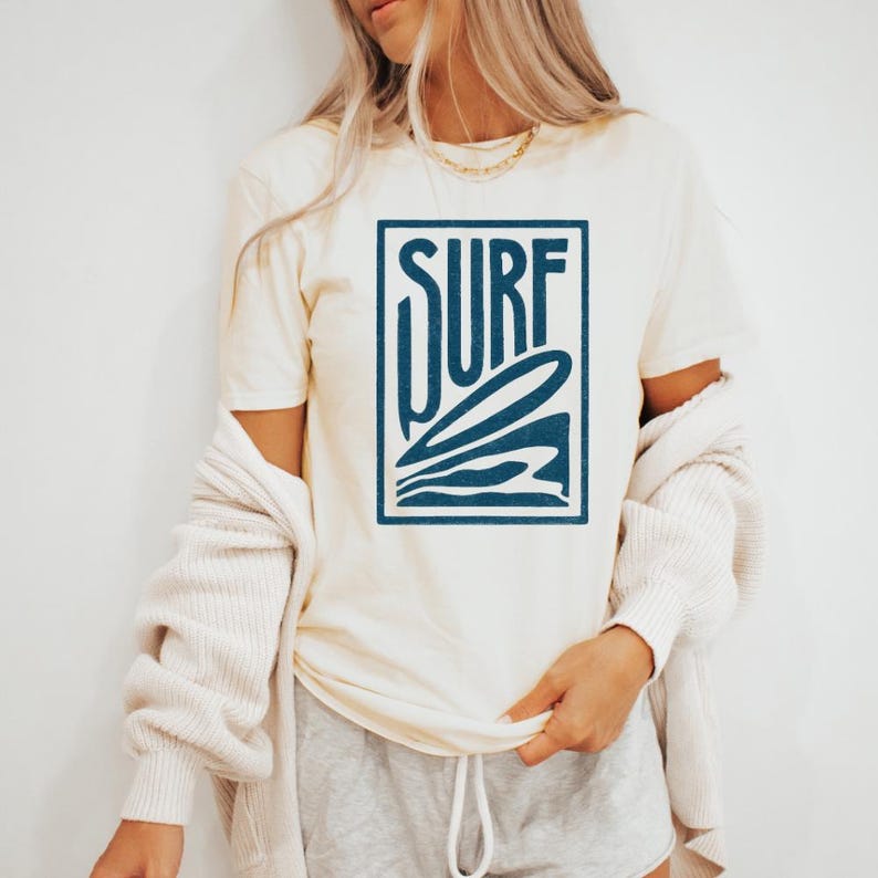 May include: A white t-shirt with a blue graphic of a surfboard and the word "SURF" in a box.