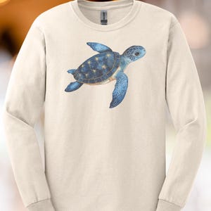 Könnte beinhalten: Cremefarbenes Langarmshirt mit einem blauen Meeresschildkröten-Design. Die Schildkröte hat einen dunkelblauen Panzer mit einem Sternbildmuster und hellere blaue Flossen. Das Shirt ist aus weichem Material.