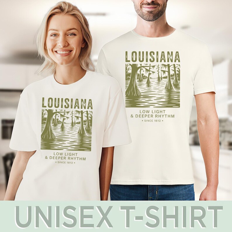 May include: Off-white unisex t-shirts featuring a Louisiana swamp scene with the text "LOUISIANA" and "LOW LIGHT & DEEPER RHYTHM SINCE 1812." The design is in olive green. The shirts are worn by two people.