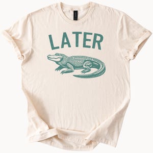 Könnte beinhalten: Ein beiges T-Shirt mit einem grünen Alligator-Motiv und dem Wort "LATER" in Grün.