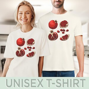 May include: Off-white unisex t-shirts featuring a detailed illustration of pomegranates. The design includes whole and sliced pomegranates in shades of red and brown. The text "UNISEX T-SHIRT" is at the bottom.