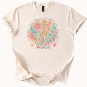 Może przedstawiać: Kremowy t-shirt z kolorowym wzorem muszli. Muszla wypełniona jest pastelowymi kwiatami, liśćmi i sercami w odcieniach różu, żółci i zieleni. Koszulka ma krótkie rękawy i okrągły dekolt.