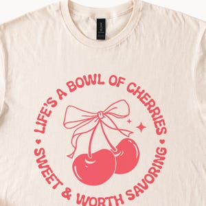 May include: A cream colored t-shirt with a pink graphic that says "Life's a bowl of cherries - sweet & worth savoring". The graphic features a cherry with a bow on top.
