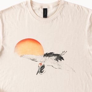 May include: A beige t-shirt with a graphic print of a crane flying in front of a red and orange sun.