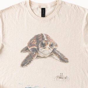 May include: A cream-colored t-shirt featuring a detailed illustration of a sea turtle in shades of brown and tan. Below the turtle, a small crab is depicted in a light brown sketch style. The shirt has a crew neckline and a black brand label.