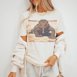 May include: Cream-colored t-shirt featuring a detailed illustration of a lizard with its tongue extended. The lizard is brown and tan, set against a desert landscape. The shirt has short sleeves and a relaxed fit.