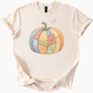 May include: Cream-colored t-shirt featuring a colorful, patchwork-style pumpkin design. The pumpkin has sections of various colors and patterns, including green, yellow, orange, and purple, with a green stem and a curling vine.