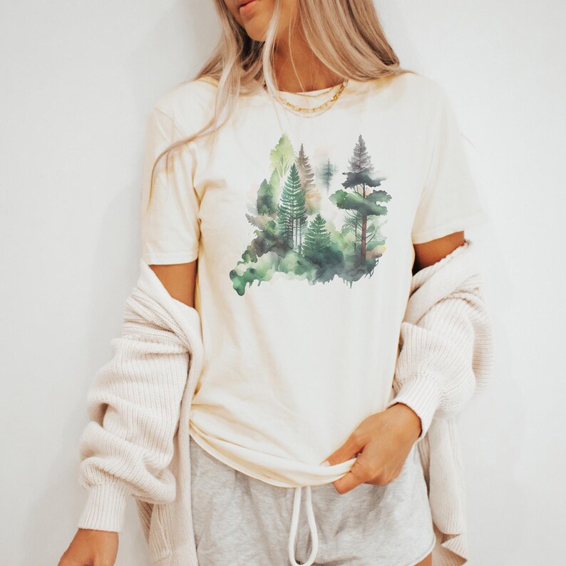 May include: A cream colored t-shirt with a watercolor design of a forest with green trees.