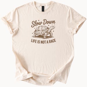 May include: Cream-coloured t-shirt featuring a brown tortoise graphic and the text "Slow Down" and "Life is Not a Race". The shirt has a crew neck and short sleeves.