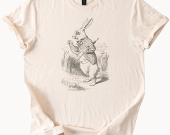 May include: Cream-colored t-shirt featuring a black and white illustration of a rabbit dressed in a waistcoat, holding a pocket watch and a walking stick. The rabbit is standing in tall grass. The shirt has a crew neck and short sleeves.