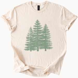 May include: A cream-colored short-sleeve t-shirt featuring a centered graphic of three evergreen trees in a muted sage green color. The tree illustration has a distressed, textured appearance, and the shirt has a classic crew neckline.