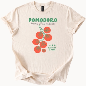 May include: Cream-colored t-shirt with a tomato graphic and text. The graphic features a cluster of red tomatoes with green stems and leaves. The text reads "POMODORO Prodotto Fresco di Napoli" and "RACCOLTO A MANO".