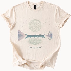 May include: Cream-colored t-shirt featuring a serene design. The graphic includes a boat on water, a floral moon, and the text "no ka ʻāina". The design is in shades of blue, green, and purple.