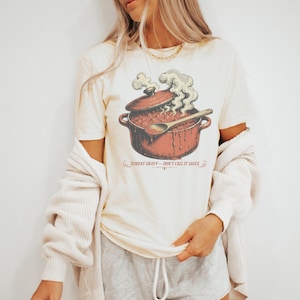 May include: A cream-colored t-shirt featuring a graphic of a red pot filled with sauce, a wooden spoon, and steam rising from the top. The text below the pot reads, "SUNDAY GRAVY - DON'T CALL IT SAUCE," with decorative flourishes on each side.