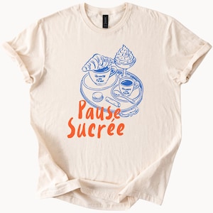 May include: Cream-colored t-shirt featuring a blue and orange graphic. The design includes a croissant, coffee, and whipped cream on a tray, with the words "Pause Sucrée" in orange.