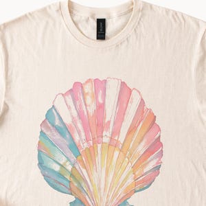 Peut inclure: Un t-shirt couleur crème arborant un grand motif central représentant un coquillage. Le coquillage est illustré dans un style aquarelle avec une palette de tons pastel doux rose, jaune, bleu et orange, présentant une texture nervurée.
