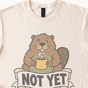 May include: A cream-colored t-shirt featuring a graphic of a brown beaver holding a yellow mug with steam rising from it. Below the beaver is a banner with the text "NOT YET" in dark gray capital letters. The design is centered on the chest.