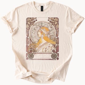 Peut inclure: T-shirt crème avec une illustration de style Art nouveau. Le motif présente le profil d'une femme aux longs cheveux blonds et coiffée d'un ornement, sur un fond circulaire orné d'éléments floraux et célestes.